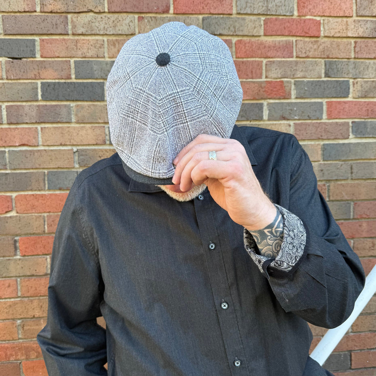 A man wearing a gray and black plaid flat cap with a black button on top, posing in front of a red and gray brick wall.