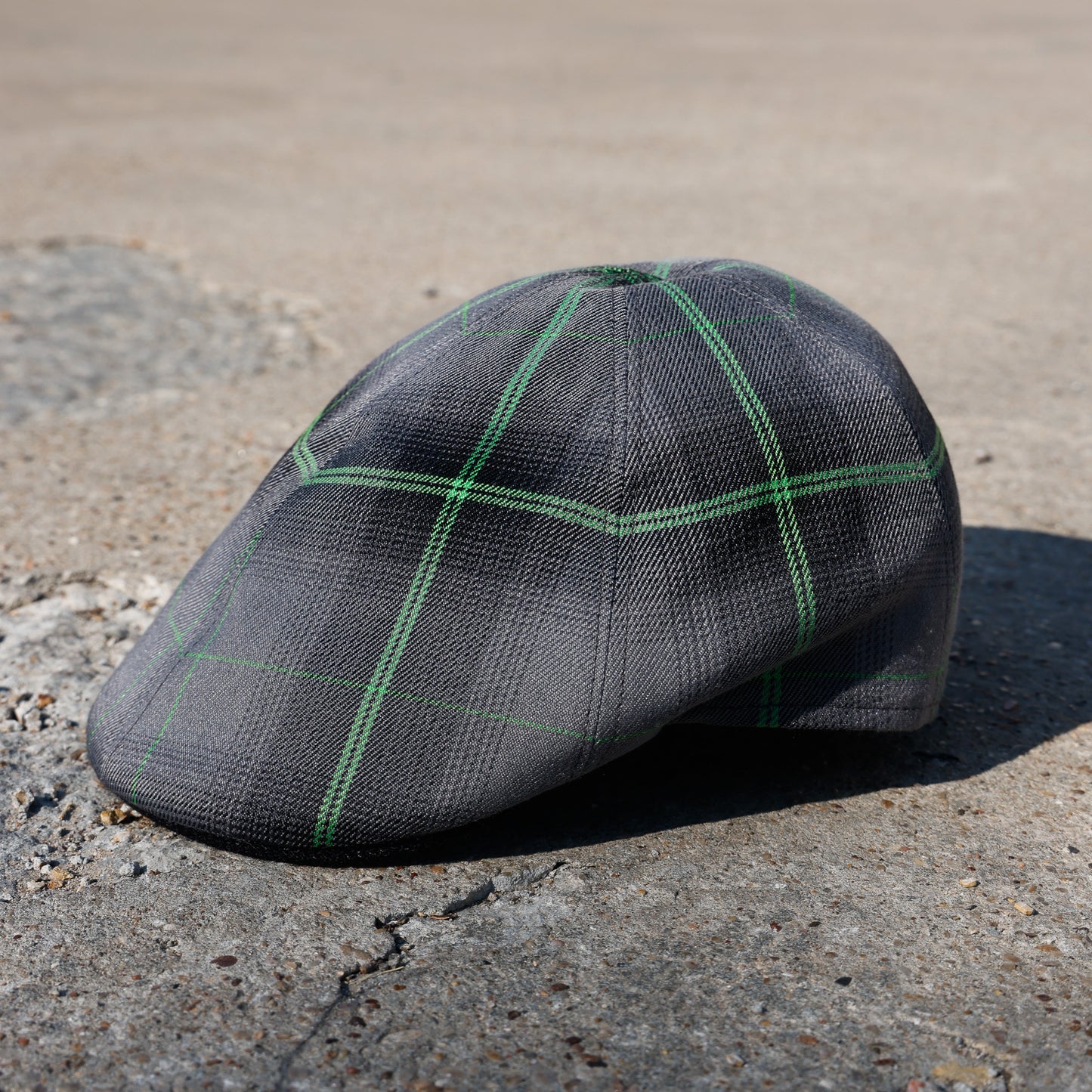 A dark gray flat cap with a green windowpane plaid pattern, resting on a textured concrete surface outdoors.