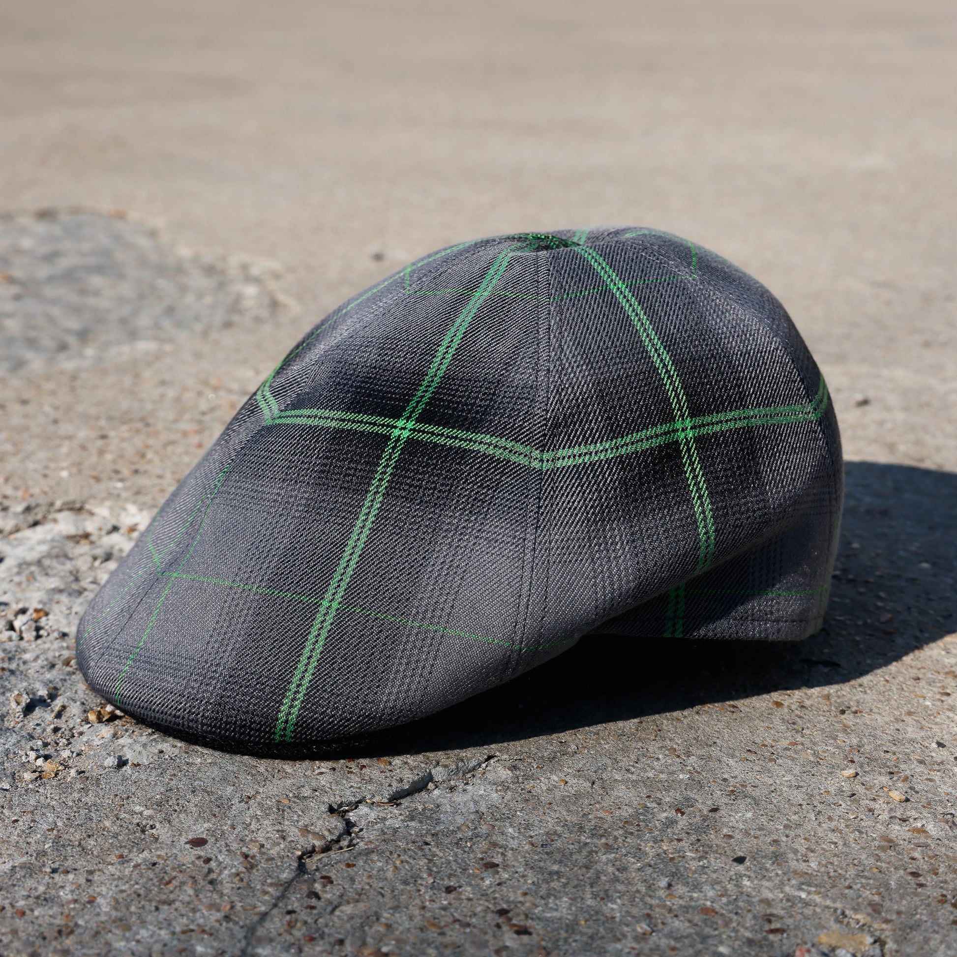 A dark gray flat cap with a green windowpane plaid pattern, resting on a textured concrete surface outdoors.
