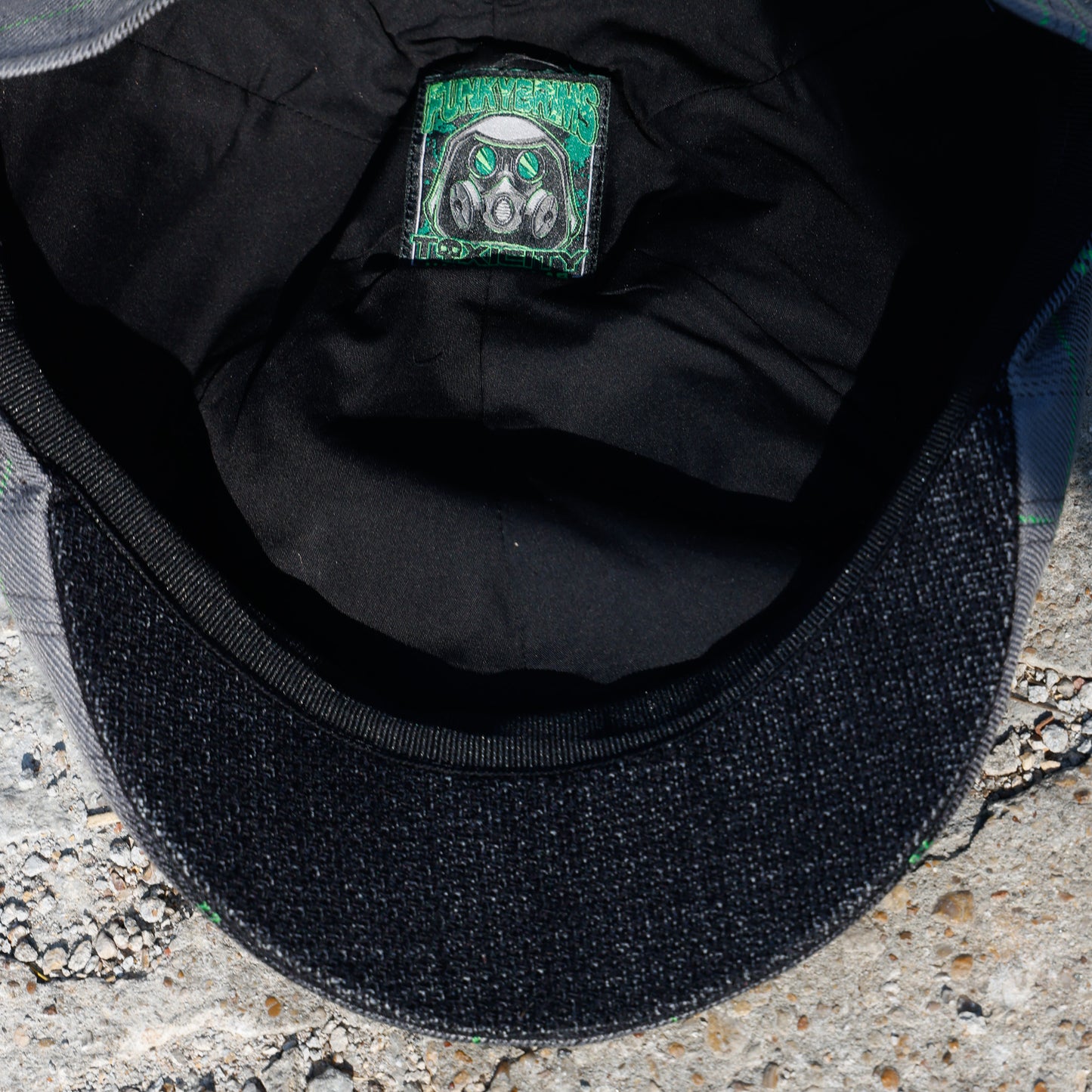 Close-up of the inside of a black baseball cap, showing a black fabric brim, black interior lining, and a square green-and-black patch with a stylized face graphic.