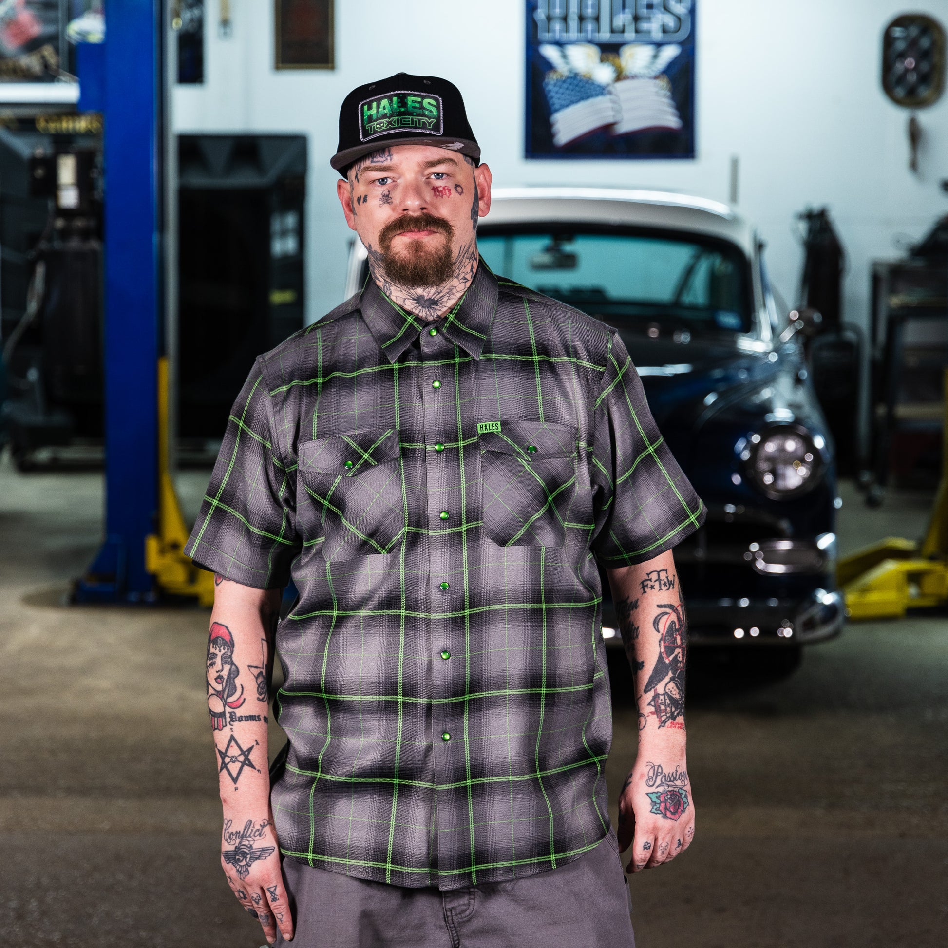 A man with tattoos stands in a garage wearing a short-sleeve gray plaid button-up shirt with green lines, a matching gray cap, and gray pants, with a blurred vintage car and workshop equipment in the background.