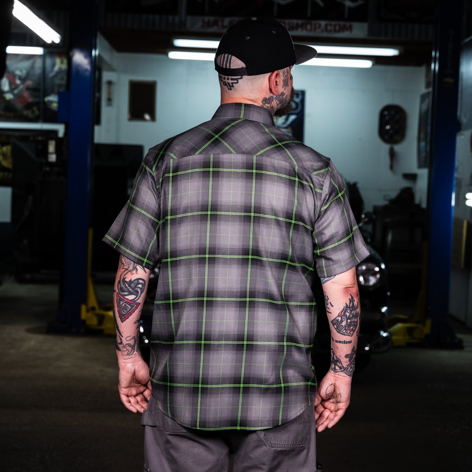 A man viewed from behind wearing a short-sleeve plaid button-up shirt in a garage setting. The shirt features a gray and black plaid pattern with bright green grid lines.