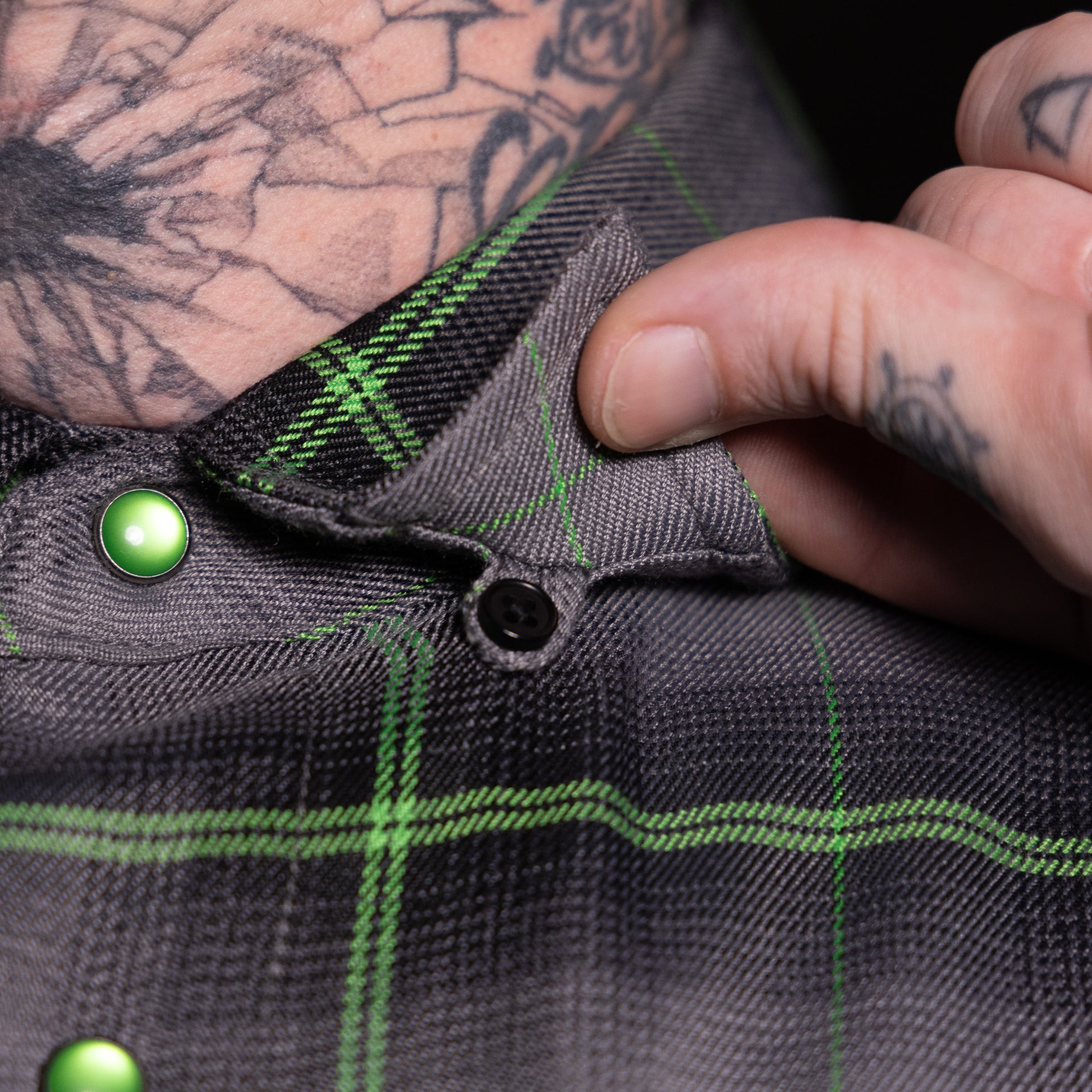 Close-up of a person wearing a dark gray textured plaid shirt with bright green lines and shiny green buttons, with a tattooed hand adjusting the collar.