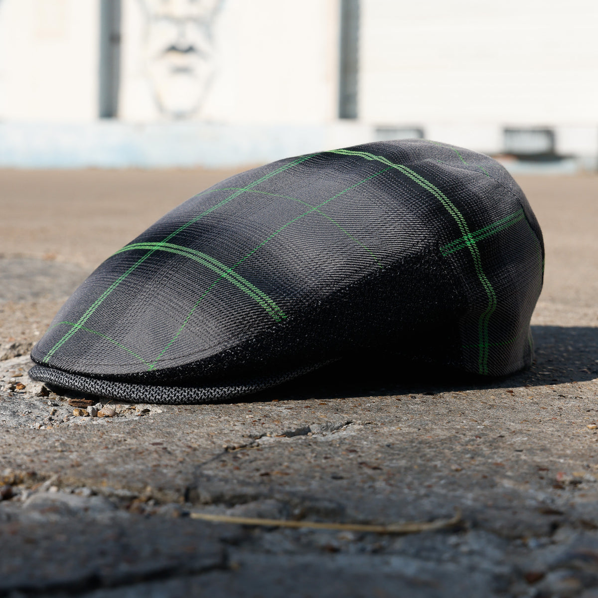 A black textured flat cap with a green plaid pattern, resting on a concrete surface outdoors.