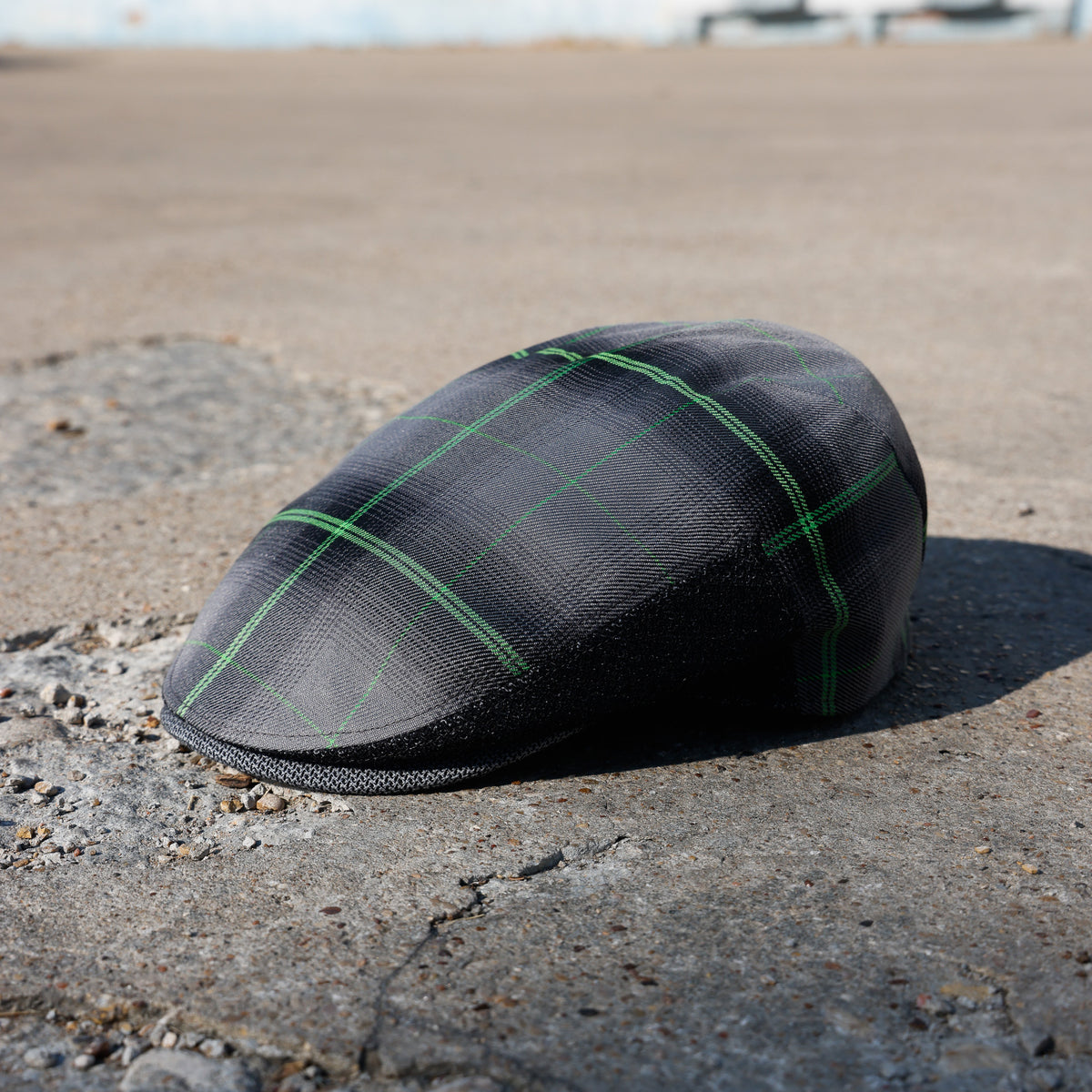 A black textured flat cap with a green plaid pattern, resting on a concrete surface outdoors.