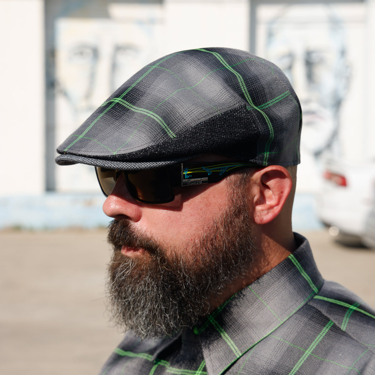 A man in profile wearing a dark gray and black plaid flat cap with green lines, matching plaid shirt, and dark sunglasses, set against a blurred outdoor background.