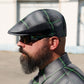 A man in profile wearing a dark gray and black plaid flat cap with bright green lines, matching plaid sunglasses, and a matching plaid collared shirt, set against an outdoor background.