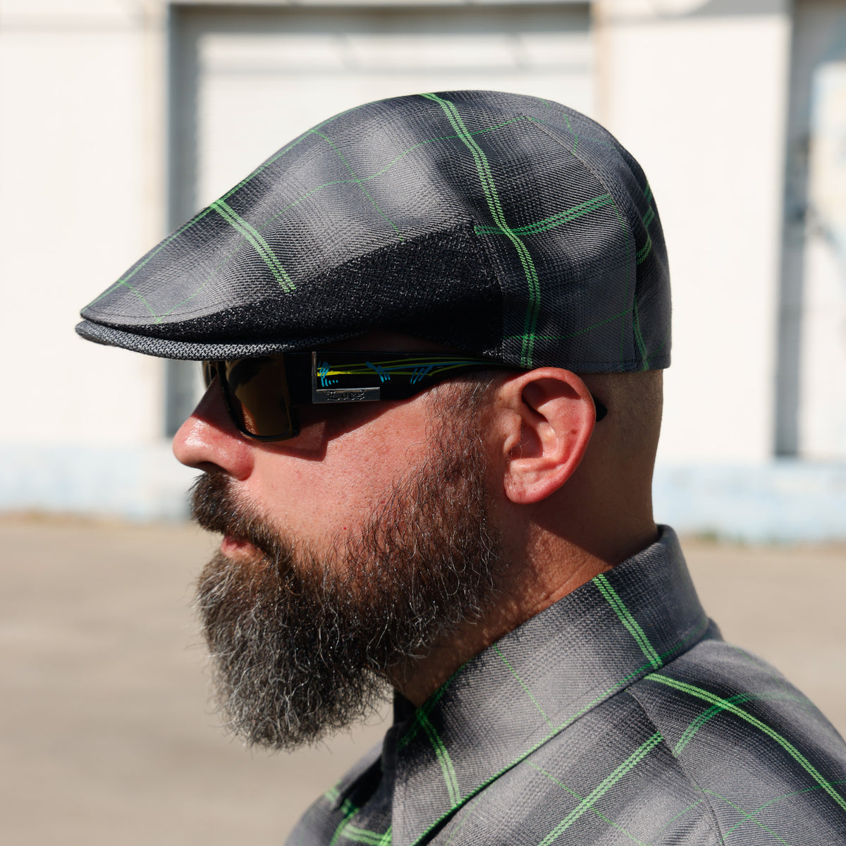 A man in profile wearing a dark gray and black plaid flat cap with bright green lines, matching plaid sunglasses, and a matching plaid collared shirt, set against an outdoor background.