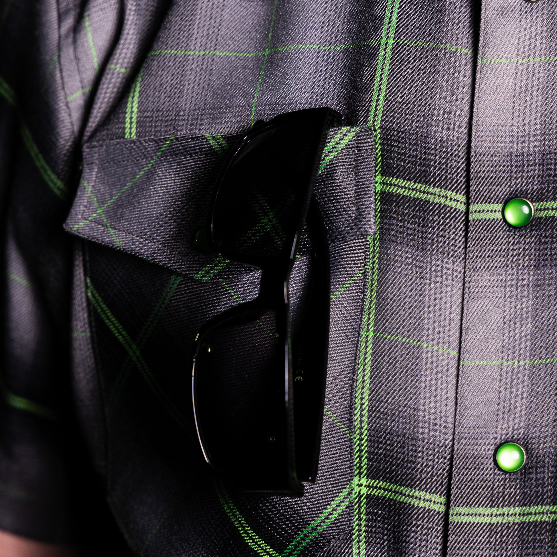 Close-up of a person wearing a dark gray plaid shirt with bright green lines and shiny green buttons, with black sunglasses tucked into the chest pocket.