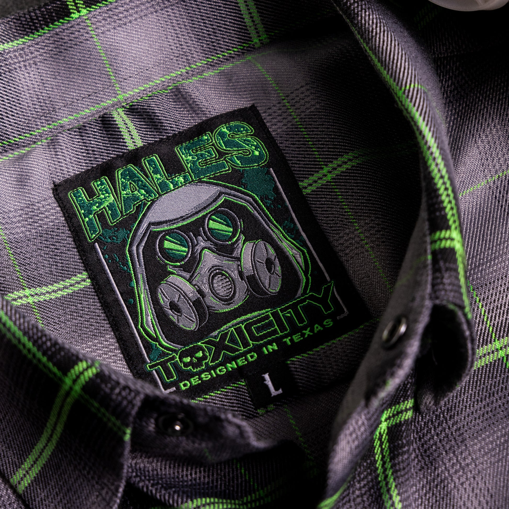Close-up of a dark gray and black plaid button-down shirt with bright green stripes, featuring a sewn-in embroidered patch with a gas mask graphic and the text 'HALES TOXICITY DESIGNED IN TEXAS'.