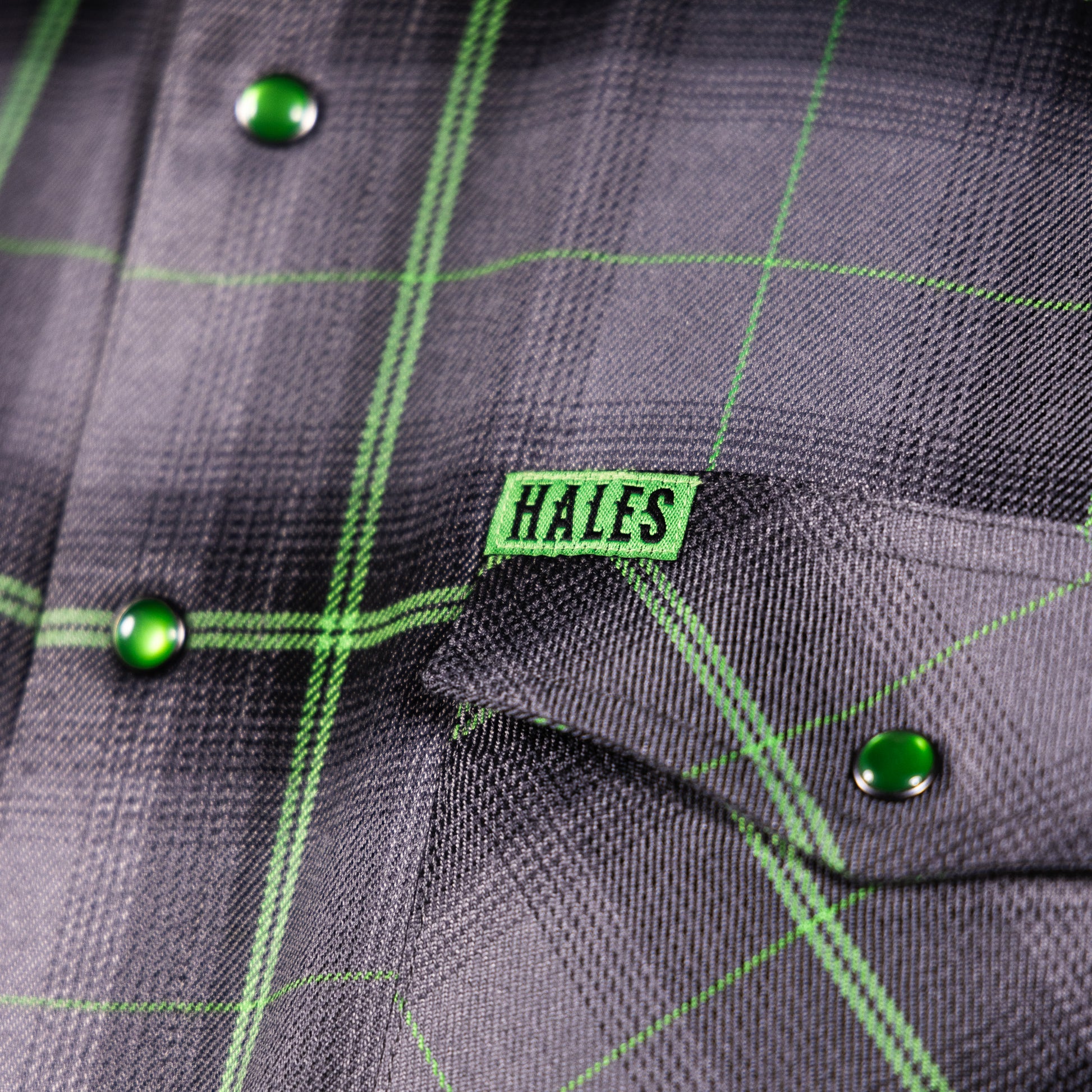 Close-up of a dark gray plaid button-up shirt with bright green windowpane lines, green buttons, and a green chest pocket flap featuring a visible Hales brand tab.