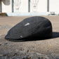 A dark charcoal gray wool flat cap with distressed details, resting on a concrete ground with a blurred urban background.