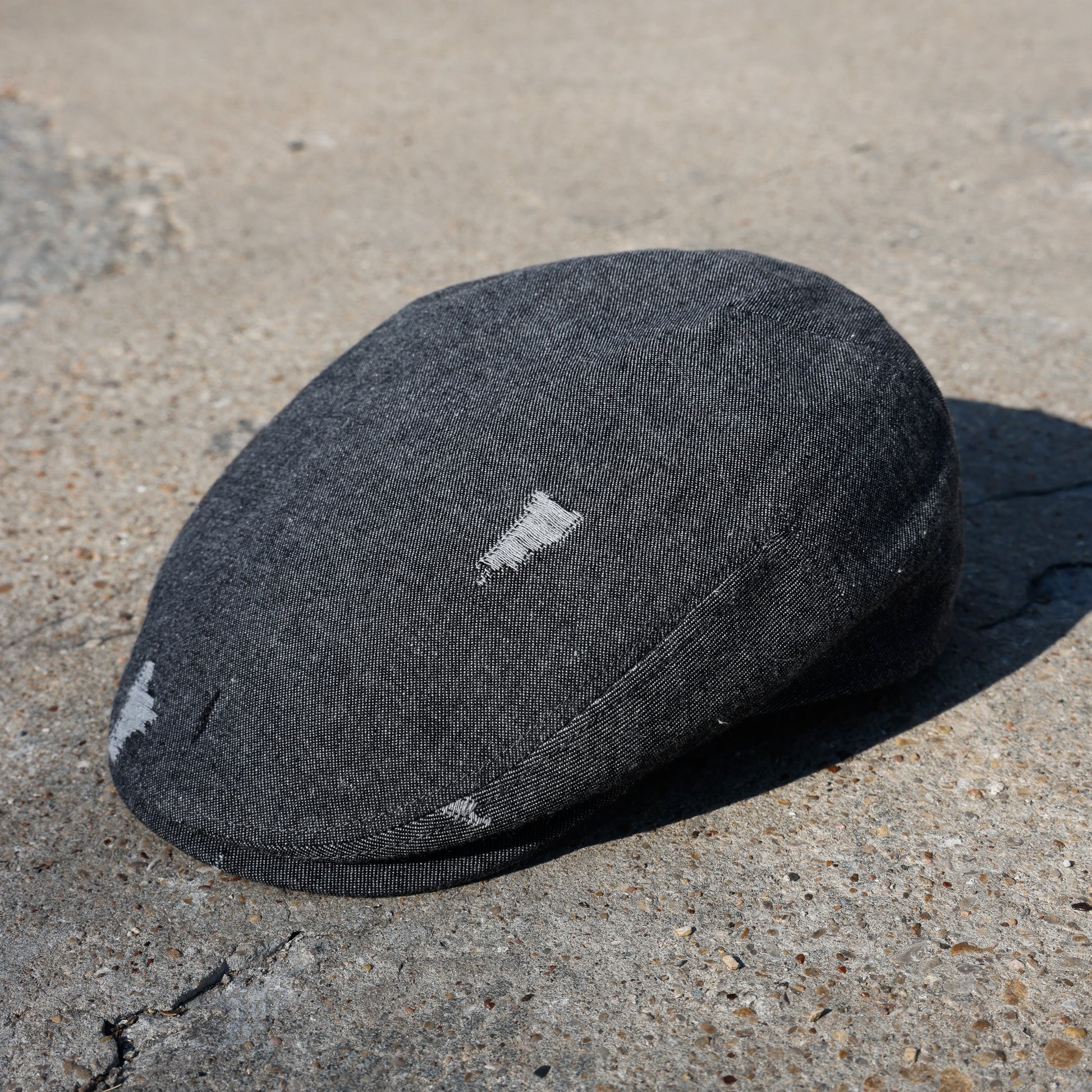 A dark charcoal gray wool flat cap with distressed white patches, resting on a textured concrete surface.
