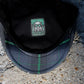 Close-up of the inside of a dark gray plaid flat cap resting on concrete, showing a green and black patch with a gas mask design.