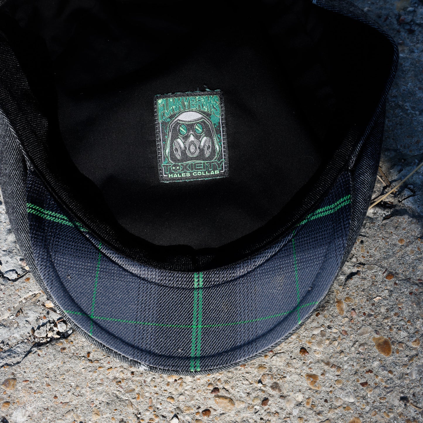 Close-up of the inside of a dark gray plaid flat cap resting on concrete, showing a green and black patch with a gas mask design.