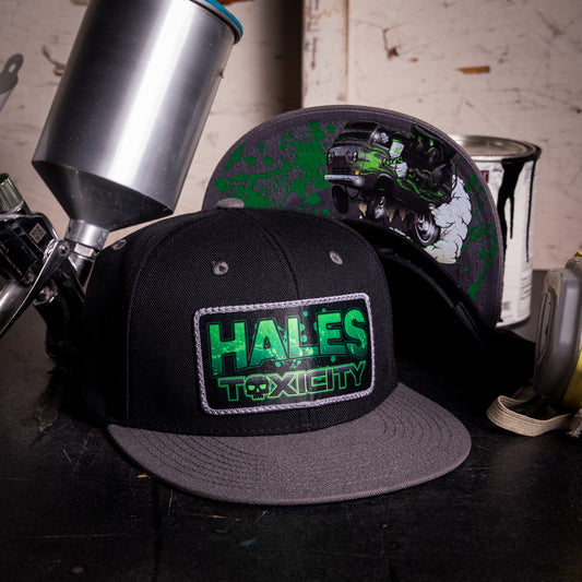 A black flat-brim cap with a gray brim and a green 'HALES TOXICITY' patch on the front, displayed alongside another cap featuring a green splatter design and a truck graphic. Spray paint cans and a paint gun are visible in the workshop-like background.