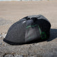 A dark gray and black patchwork flat cap with a green embroidered logo, resting on a concrete surface outdoors.