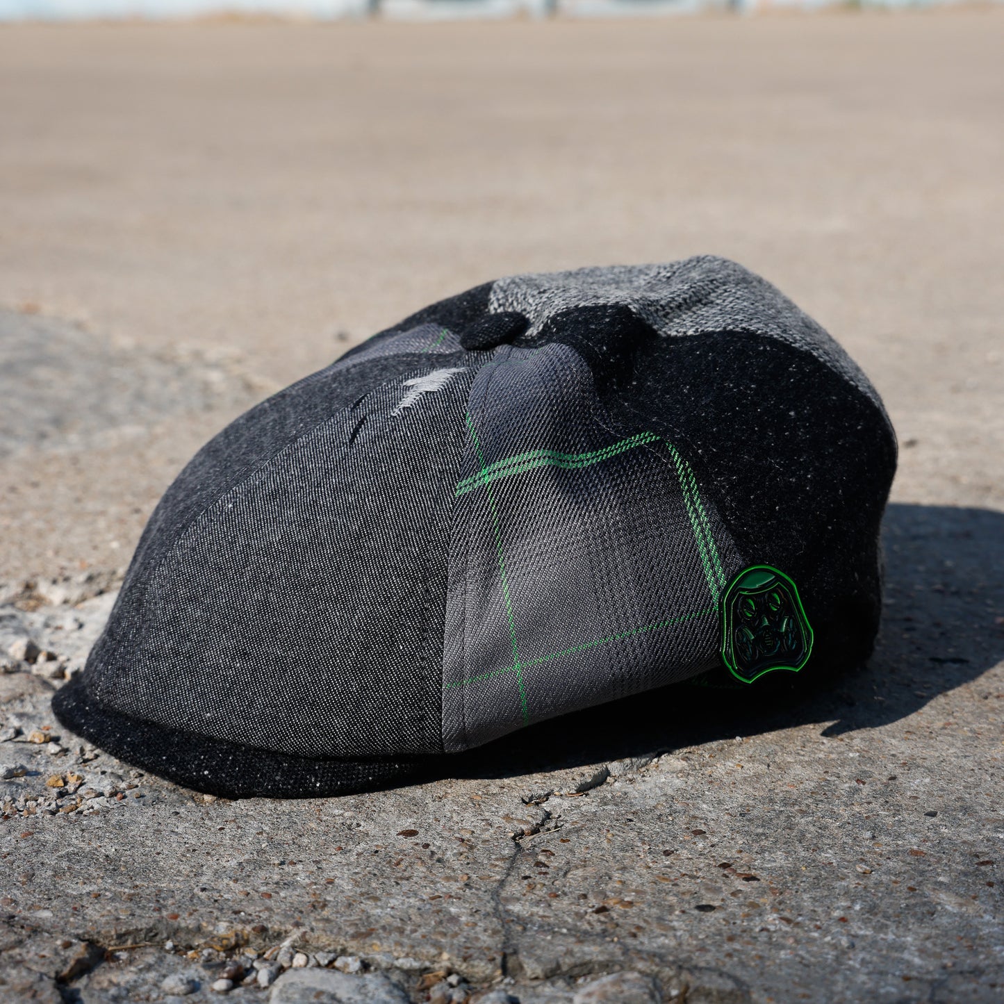 A dark gray and black patchwork flat cap with a green embroidered logo, resting on a concrete surface outdoors.