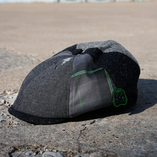 A dark gray and black patchwork flat cap with a green embroidered logo, resting on a concrete surface outdoors.