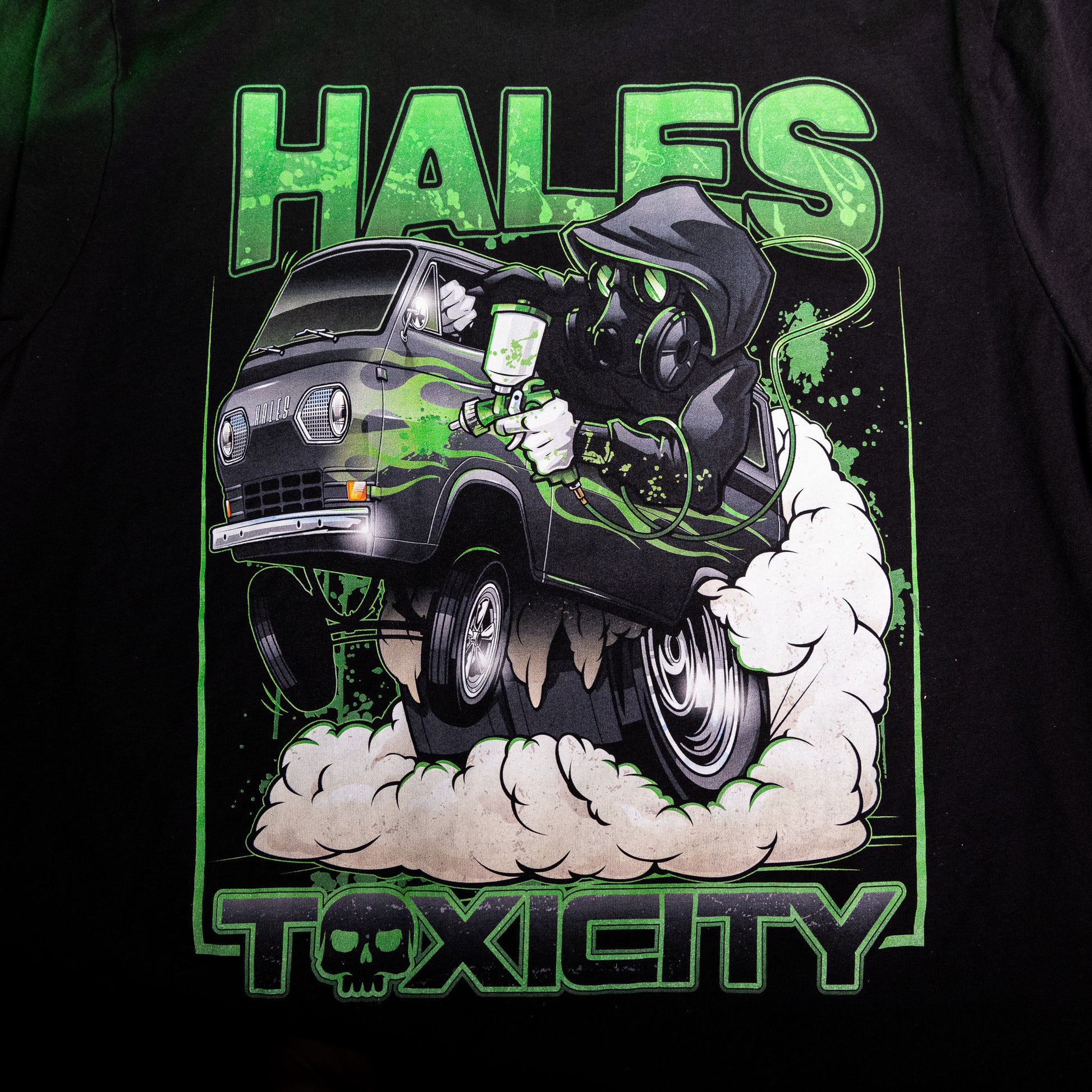 Close-up of a black graphic t-shirt featuring a green and white illustration of a hooded figure driving a van with smoke, with the text 'HALES' at the top and 'TOXICITY' at the bottom.