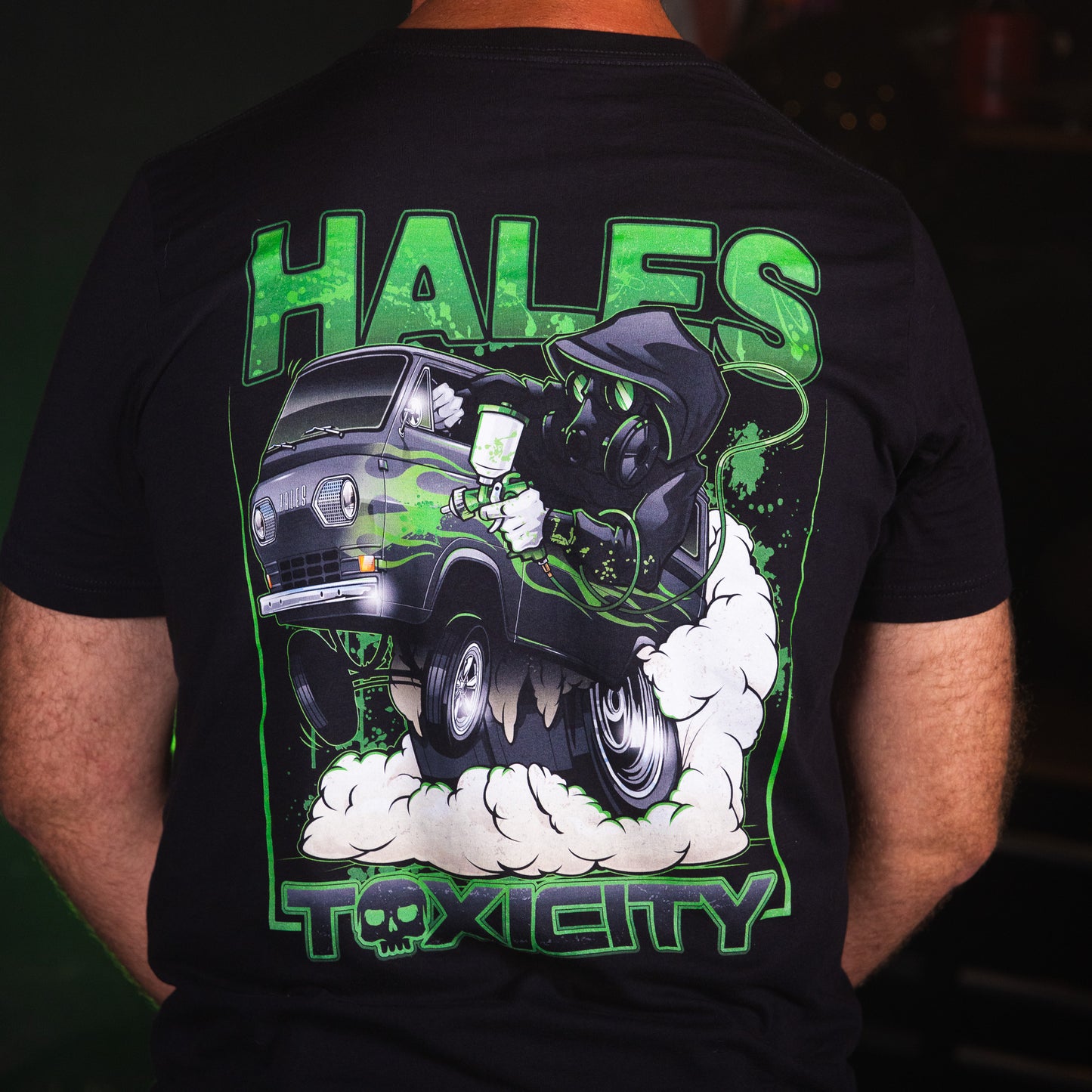 A person shown from the back wearing a black graphic T-shirt. The shirt features a large green, white, and gray illustration of a hooded figure in a van with the words 'HALES' at the top and 'TOXICITY' at the bottom.