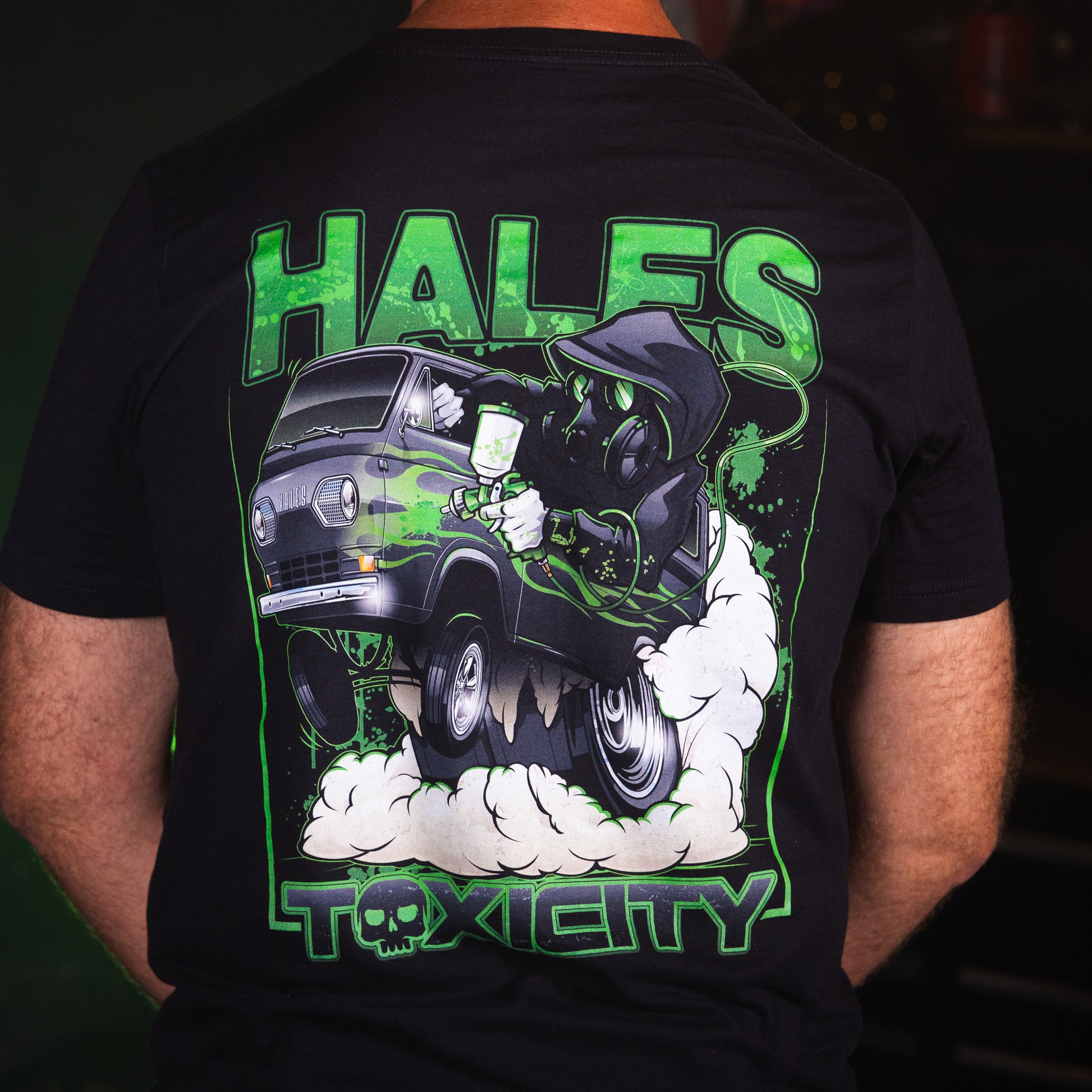 A person shown from the back wearing a black graphic T-shirt. The shirt features a large green, white, and gray illustration of a hooded figure in a van with the words 'HALES' at the top and 'TOXICITY' at the bottom.
