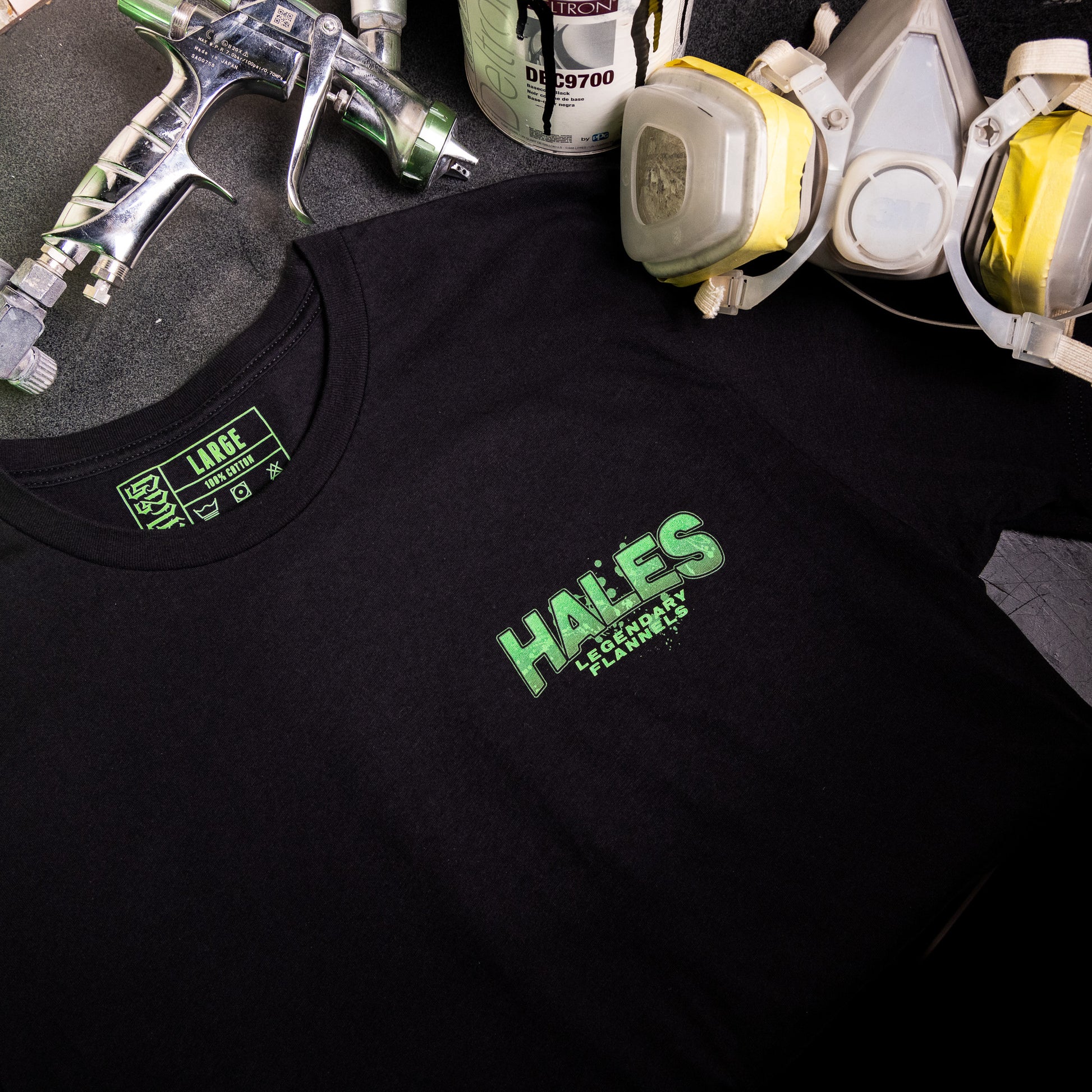 A black short-sleeve t-shirt with a green 'HALES' graphic on the chest, styled with spray paint equipment and paint cans on a dark surface.