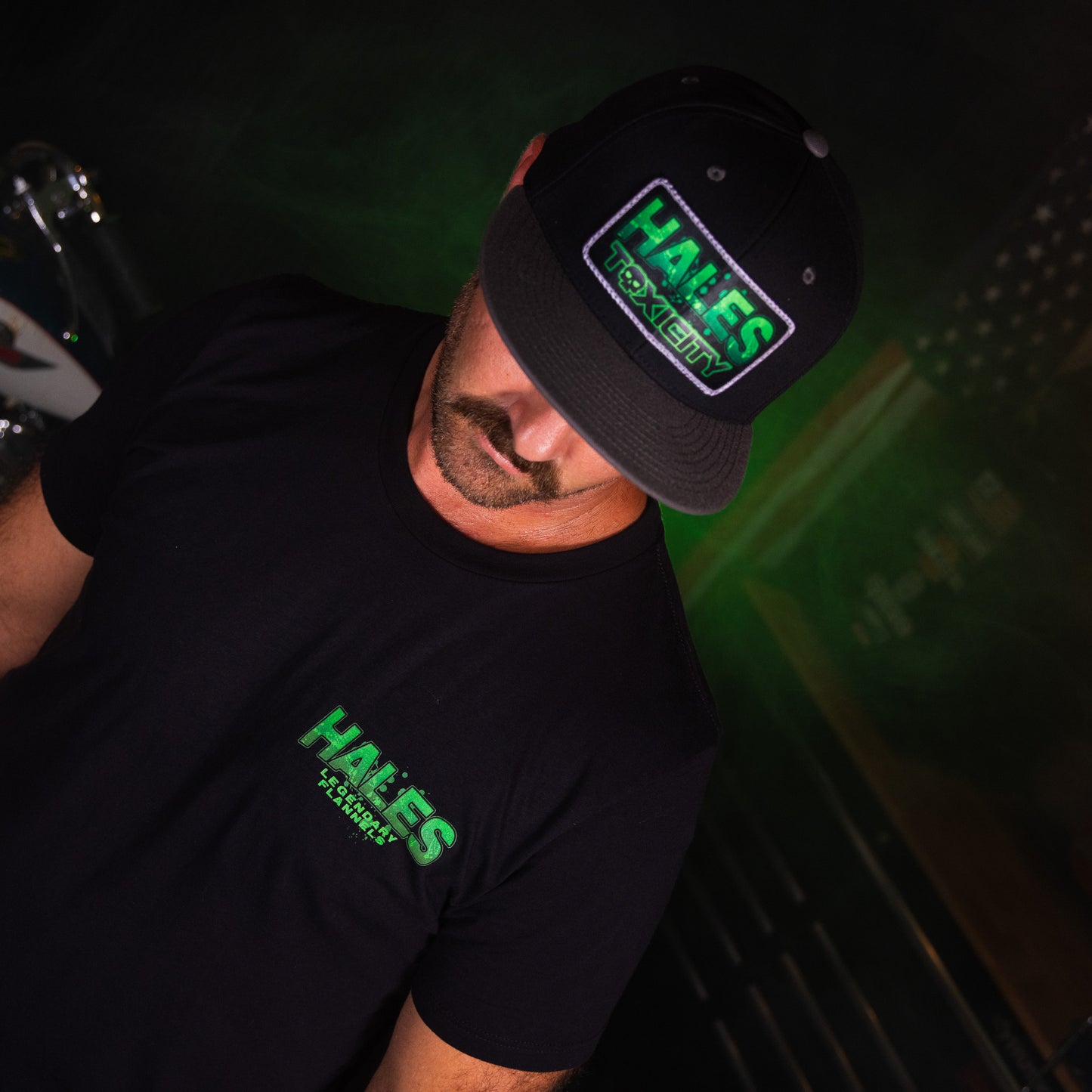A man wearing a black t-shirt and a black baseball cap, both featuring a green and white 'HALES TOXICITY' graphic print, in a dark, moody setting with green ambient lighting.
