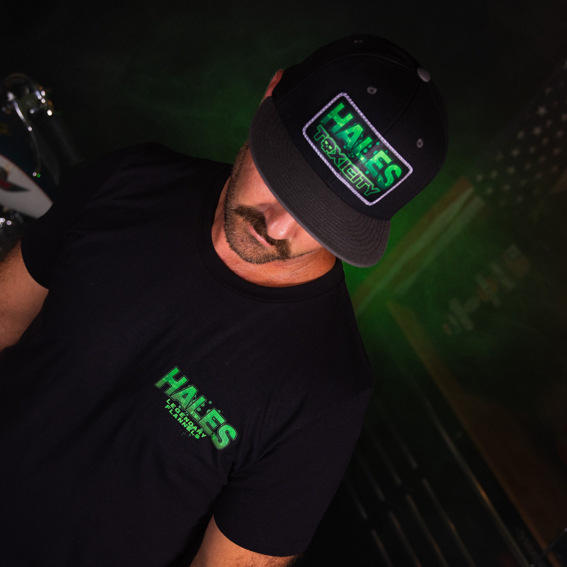 A man wearing a black t-shirt and a black baseball cap, both featuring a green and white 'HALES TOXICITY' graphic print, in a dark, moody setting with green ambient lighting.