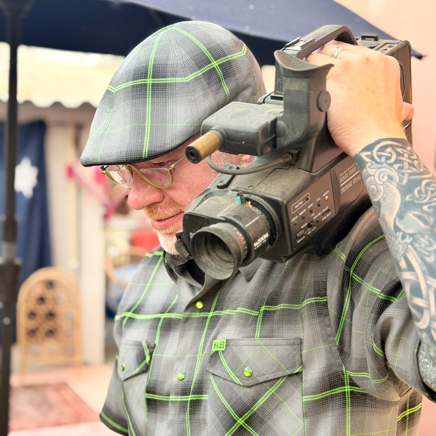 A man wearing a gray plaid flat cap and matching gray plaid button-up shirt with bright green grid lines, holding a vintage black video camera over his shoulder in an outdoor setting.