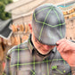 A man wearing a gray and black plaid flat cap with bright green lines, paired with a matching short-sleeve button-down shirt. He is adjusting the brim of the cap.