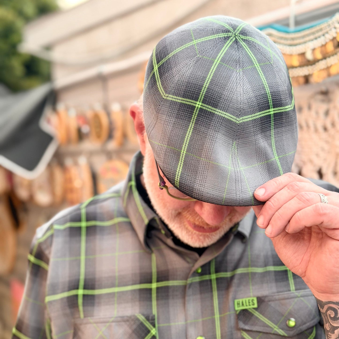 A man wearing a gray and black plaid flat cap with bright green lines, paired with a matching short-sleeve button-down shirt. He is adjusting the brim of the cap.