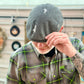 A man wearing a gray distressed flat cap and a green and brown plaid button-down shirt, holding the brim of the cap with his hand.