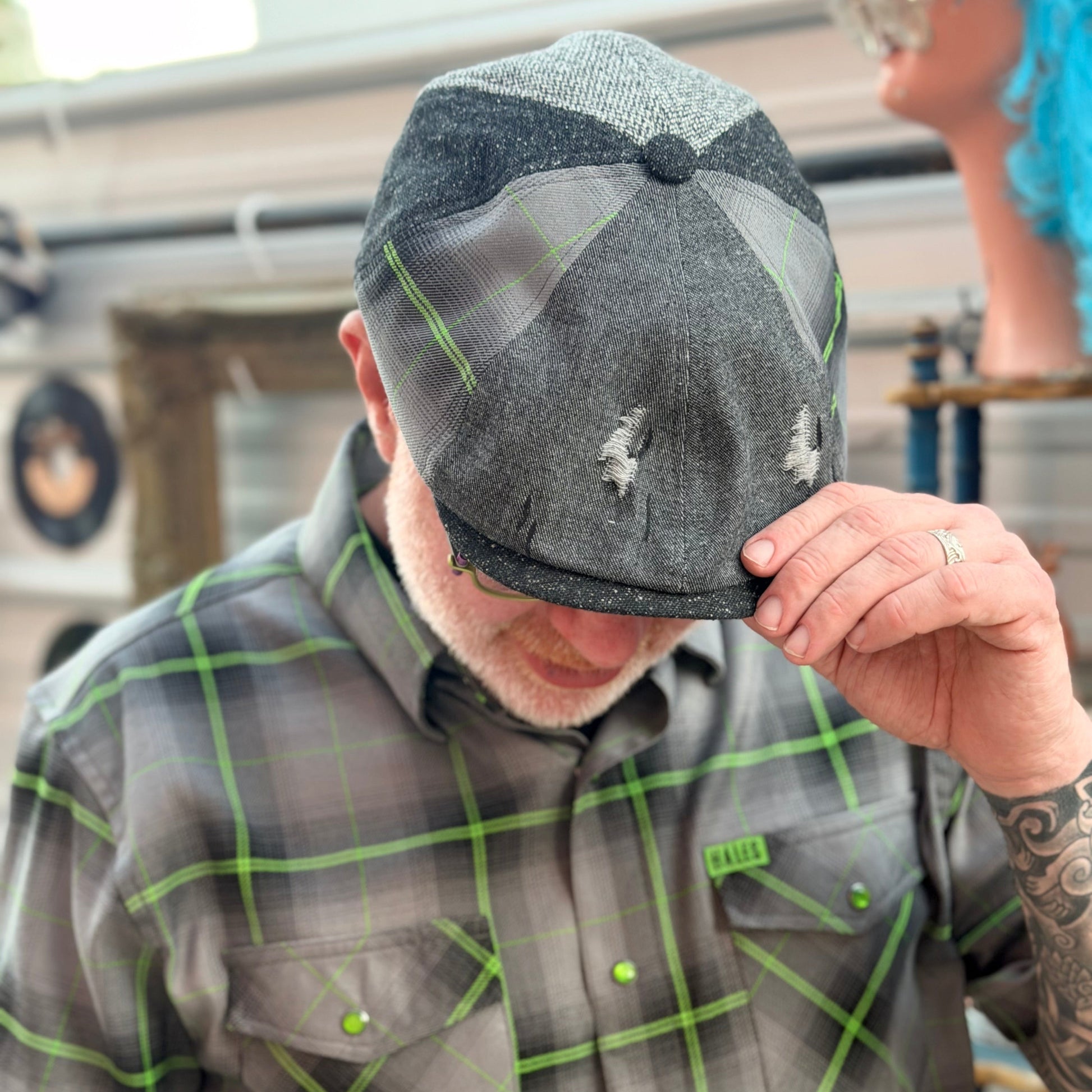 A man wearing a dark gray and black distressed flat cap and a matching gray plaid short-sleeve button-up shirt with neon green accents, holding the brim of the cap with his hand.