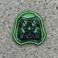 A green and black enamel pin shaped like a gas mask helmet, resting on a wooden surface.
