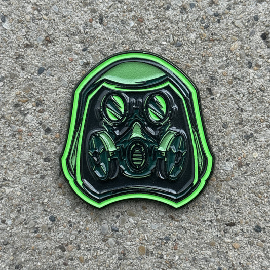 A green and black enamel pin shaped like a gas mask helmet, resting on a wooden surface.