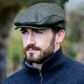 A man wearing a textured herringbone flat cap in olive green and brown, paired with a dark navy ribbed zip-up sweater, set against a soft, blurred outdoor background.