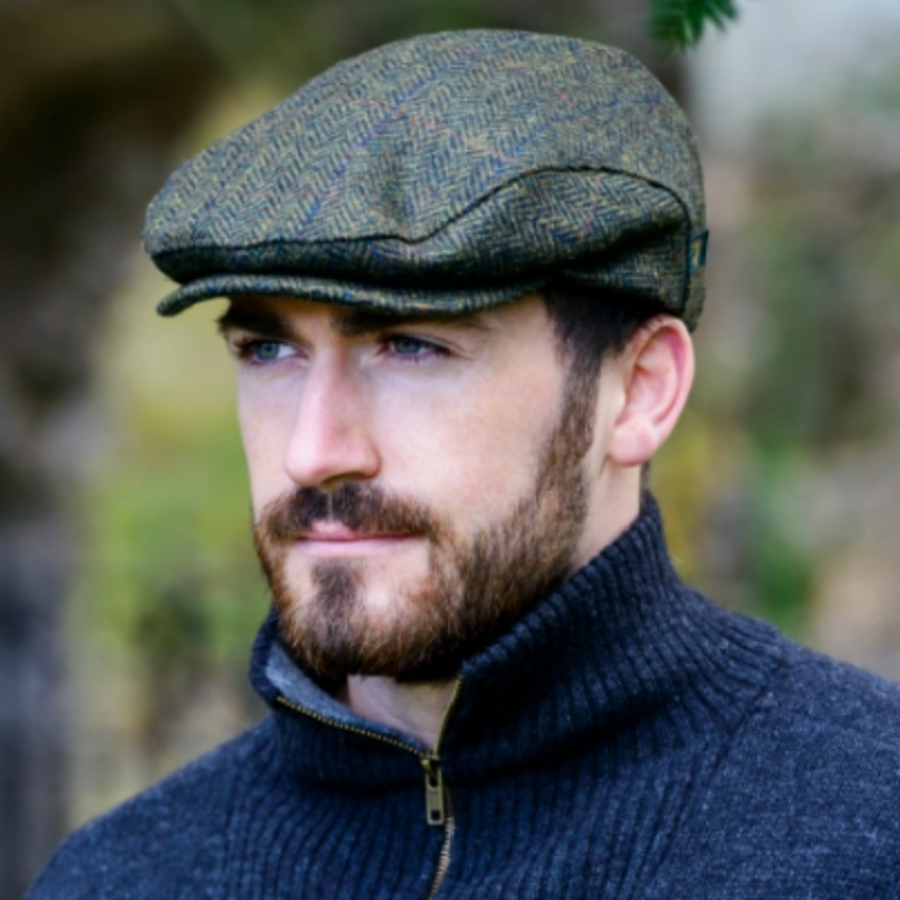 A man wearing a textured herringbone flat cap in olive green and brown, paired with a dark navy ribbed zip-up sweater, set against a soft, blurred outdoor background.
