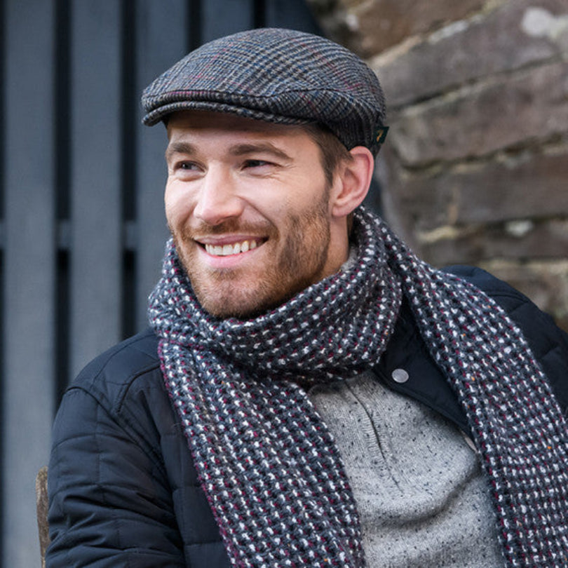 Man wearing a cap and scarf, smiling outdoors against a brick wall.