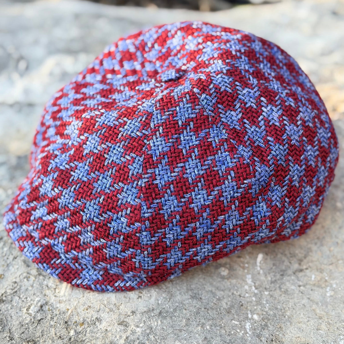 The Troubadour burgundy-red and denim-blue houndstooth newsboy cap resting on stone, front three-quarter view.