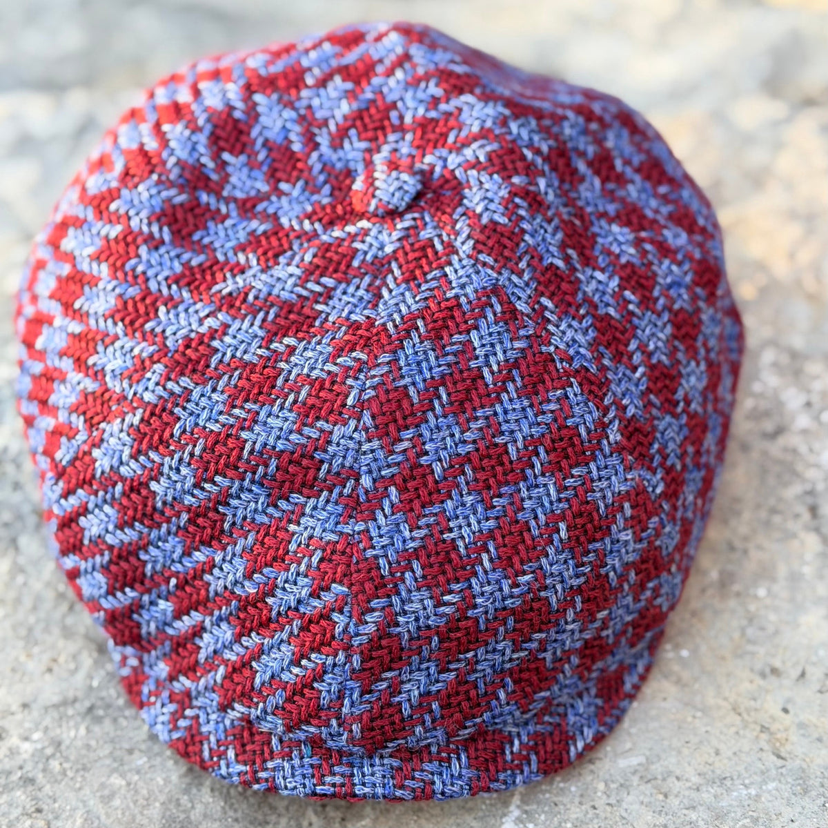 The Troubadour burgundy-red and denim-blue houndstooth newsboy cap resting on stone, top view.