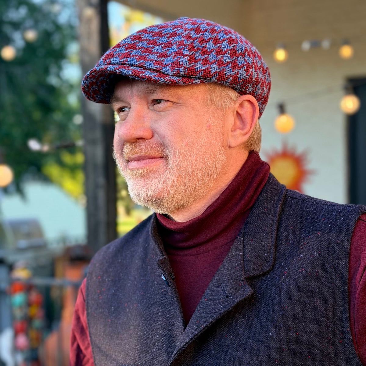The Troubadour houndstooth newsboy cap worn with a burgundy turtleneck and dark vest, three-quarter side view.