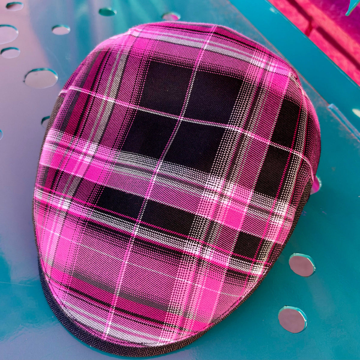 A close-up of a small oval-shaped product, likely a pouch or accessory cover, featuring a vibrant pink and black plaid pattern with thin white accent lines, resting on a teal metal surface.