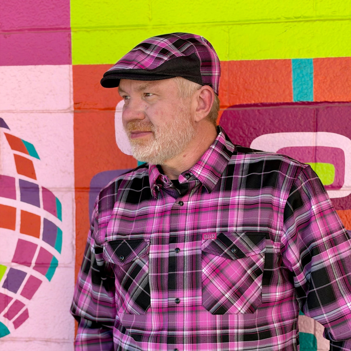 A man wearing a pink, purple, and black plaid button-down shirt and matching plaid flat cap, standing in front of a brightly colored painted brick wall.