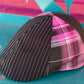 A structured flat cap with a three-panel design: one panel is black with thin white pinstripes, and two adjacent panels are pink plaid with black and white lines. The cap is photographed on a bright turquoise surface with a colorful abstract background.