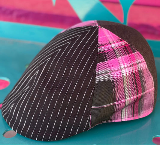 A structured flat cap with a three-panel design: one panel is black with thin white pinstripes, and two adjacent panels are pink plaid with black and white lines. The cap is photographed on a bright turquoise surface with a colorful abstract background.