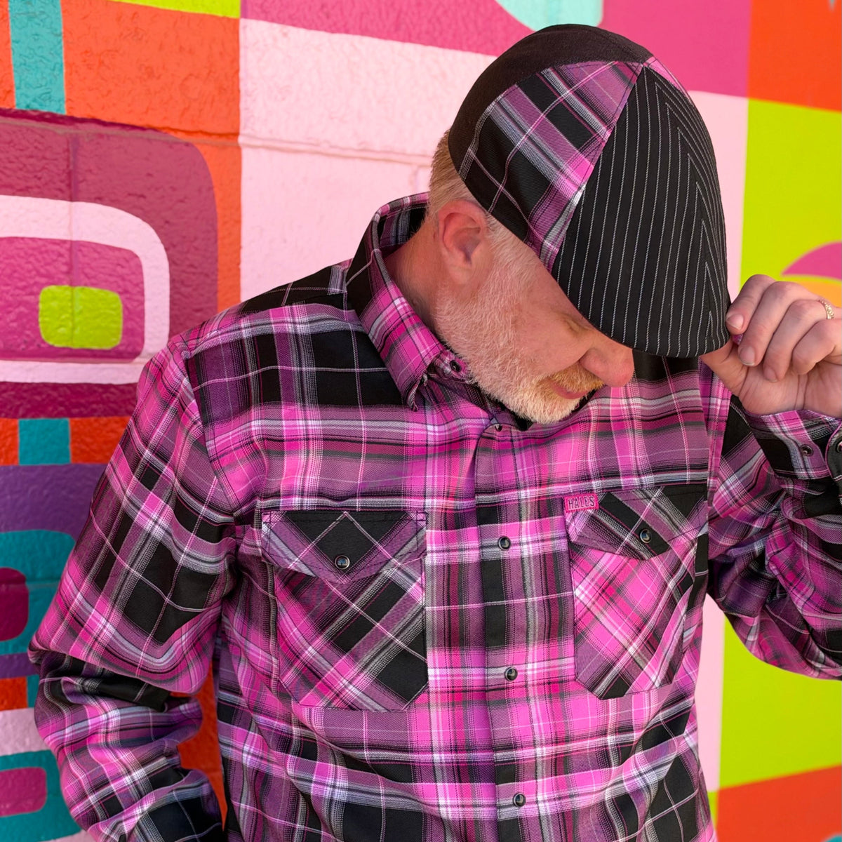 A man wearing a vibrant pink and black plaid button-down shirt and a matching pink and black plaid cap, posed in front of a colorful geometric painted brick wall.
