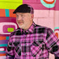 A man wearing a pink and black plaid button-down shirt and a matching black pinstripe flat cap, standing in front of a colorful graffiti wall.