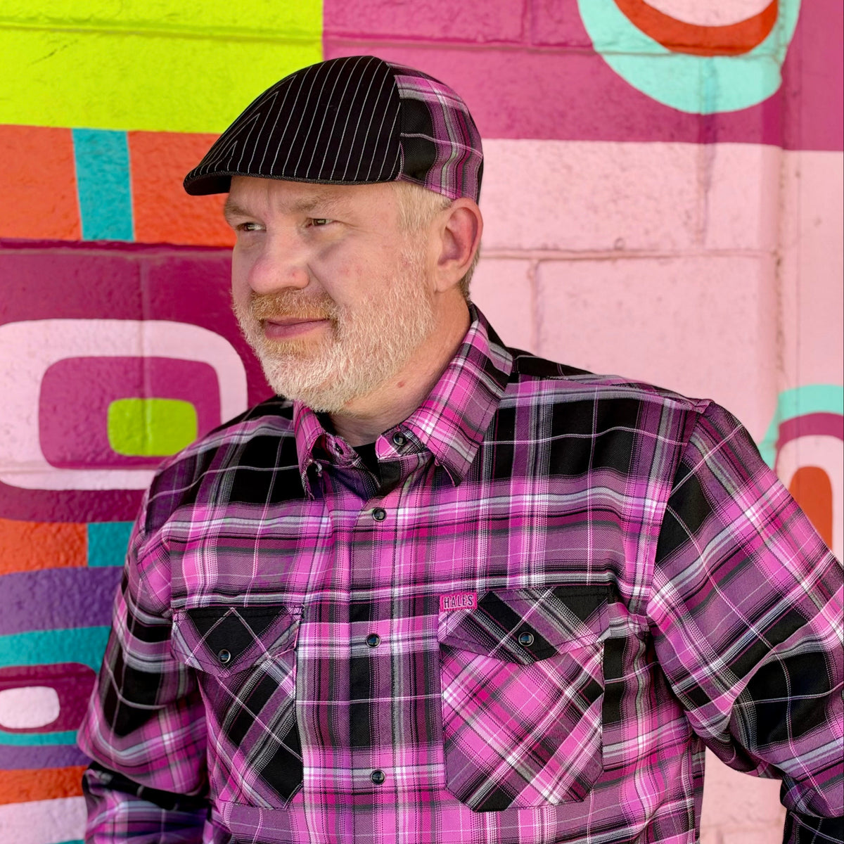A man wearing a pink and black plaid button-down shirt and a matching black pinstripe flat cap, standing in front of a colorful graffiti wall.