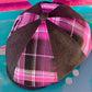 A structured flat cap with a dark brown herringbone fabric crown and bright pink, black, and white plaid panels, resting on a turquoise surface.